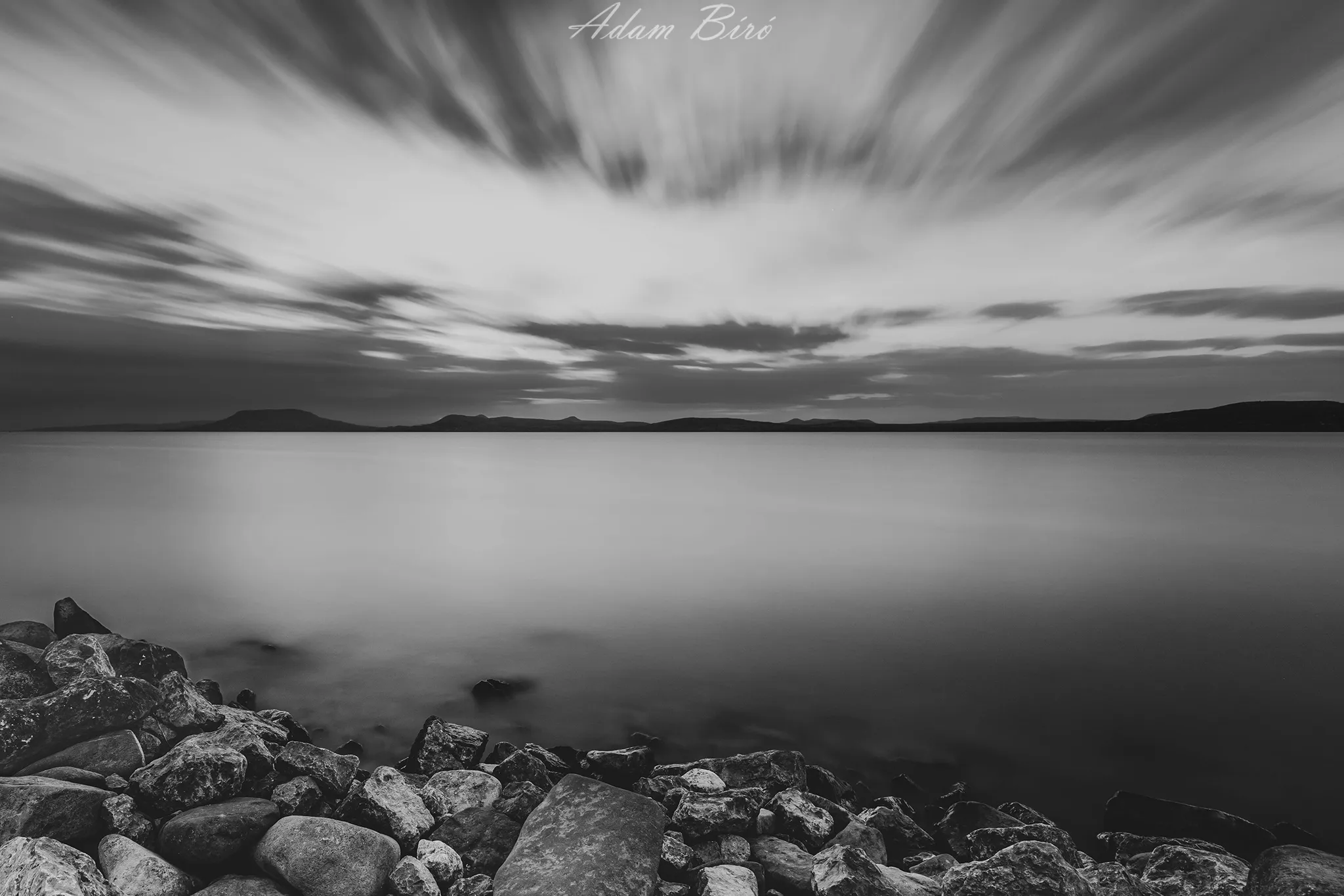 Black and white long exposure landscape at Lake Balaton with dramatic clouds, calm water and rocky foreground